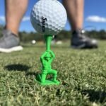 Green toy soldier golf tee holding a golf ball on grass, with sneakers behind