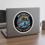 USS Charlotte Silent Stinger sticker on the lid of a gray laptop beside a coffee mug